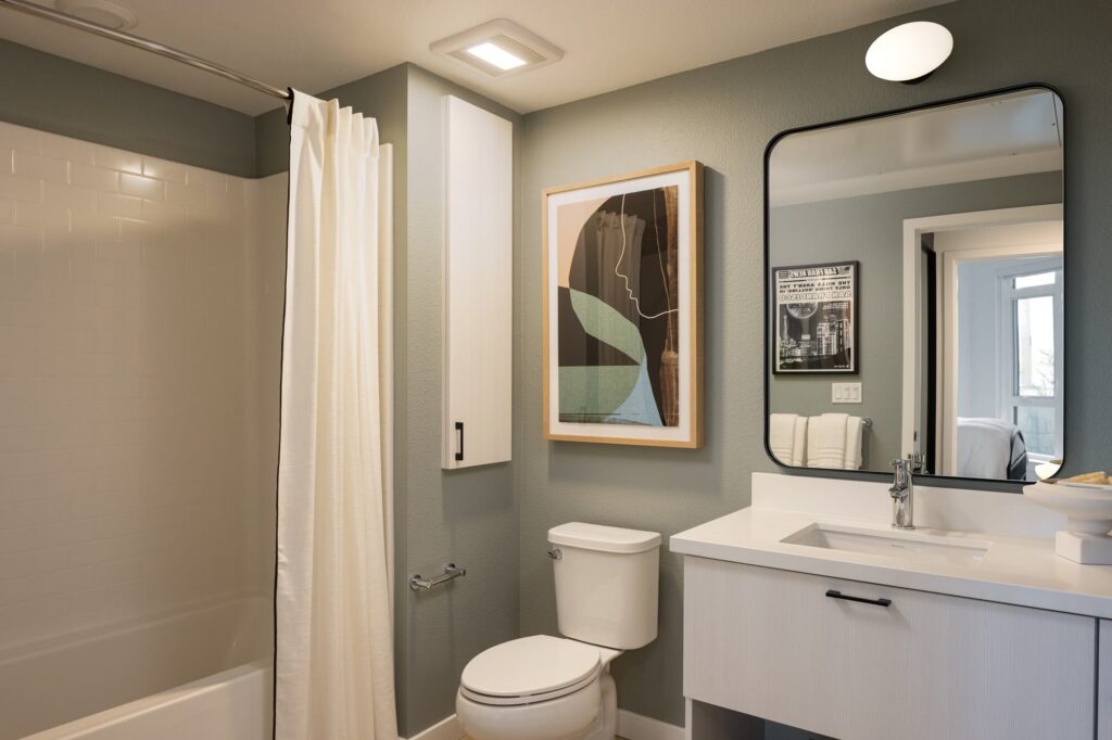 Modern bathroom with a bathtub and white shower curtain, toilet, sink with a mirror, abstract wall art, and soft green walls. A towel hangs near the window, and toiletries sit on the counter.