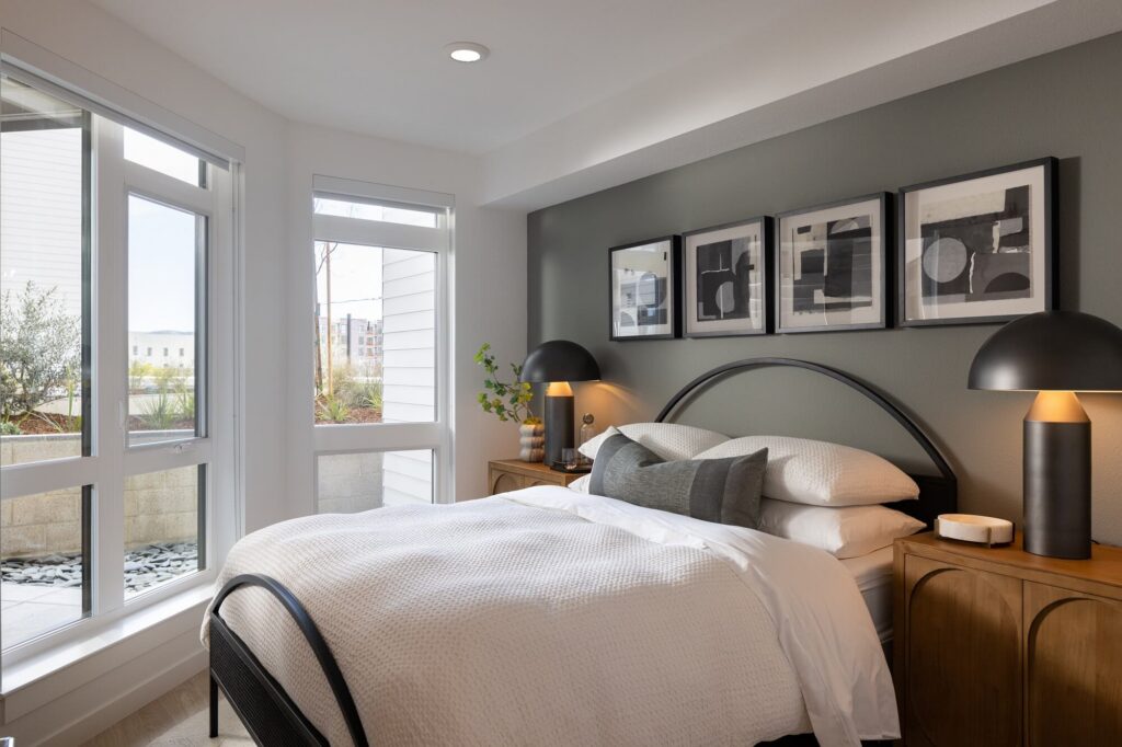 A modern bedroom with a bed, two wooden nightstands, black lamps, and abstract art on the gray accent wall. Large windows let in natural light.