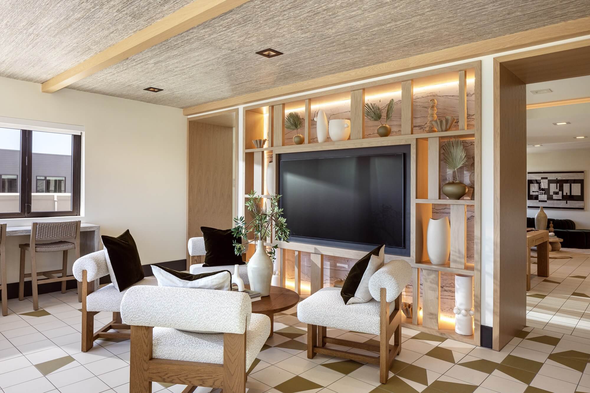 Modern lounge with cream armchairs, a round table, and built-in shelving around a large TV. Shelves display vases and plants. Window and desk are visible to the left; another seating area appears in the background.