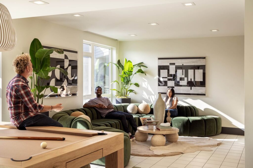 Three people relax and converse in a modern lounge with green sofas, plants, abstract art, a coffee table, and a pool table in the foreground. Sunlight streams in through large windows.