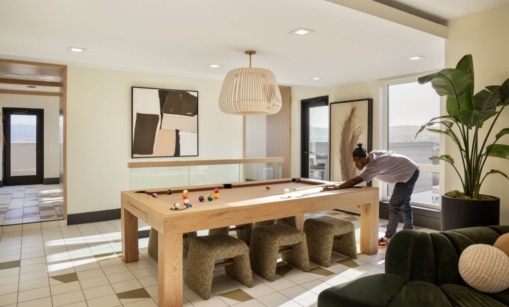 A person leans over a pool table, aiming to take a shot in a modern, sunlit room with abstract art, stools, a large plant, and tiled floors.