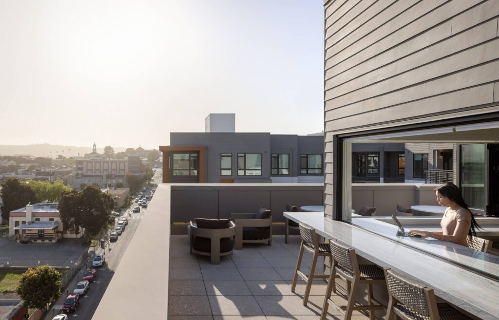 A person works on a laptop at a rooftop patio with tables and chairs, overlooking a city street and buildings in the sunlight.