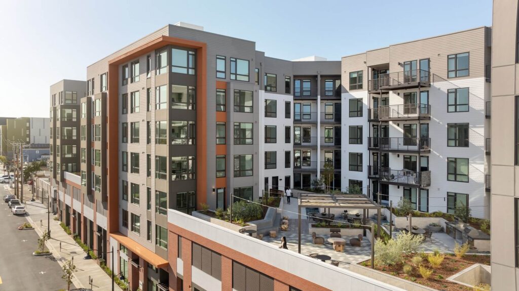 Modern multi-story apartment building with large windows, balconies, and a central courtyard featuring outdoor seating, landscaping, and walkways. The street and sidewalk are visible on the left side under a clear sky.