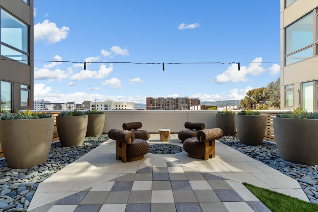 Outdoor patio with four patterned chairs around a small wooden table, large potted plants, decorative stones, string lights overhead, and buildings visible in the background under a blue sky with clouds.