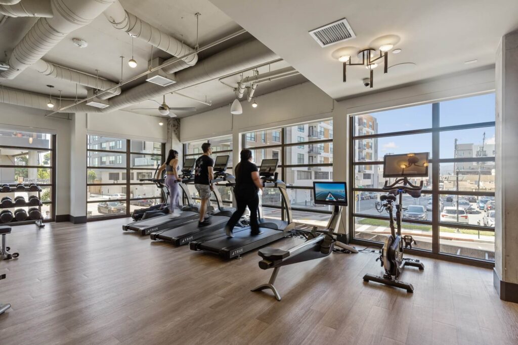 Three people are using treadmills in a modern gym with large windows, wooden floors, and a view of buildings outside. Two stationary bikes and a row of dumbbells are also visible.