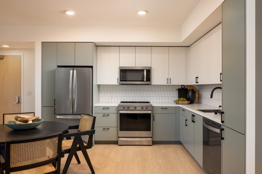Modern kitchen with gray and white cabinets, stainless steel appliances, a dining table with chairs, and a bowl of green apples on the counter. Light wood flooring and white tiled backsplash are visible.