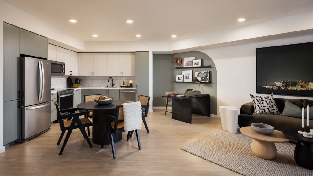 Modern open-concept kitchen and living area with stainless steel appliances, a round dining table, black chairs, a black sofa, wall art, and neutral decor. Light wood floors and recessed lighting brighten the space.