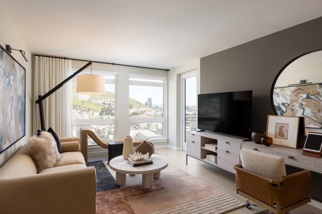 Modern living room with beige sofa, round coffee table, TV on white console, large wall art, a circular mirror, and large windows showing a city view. Neutral tones and natural light create a cozy, stylish atmosphere.
