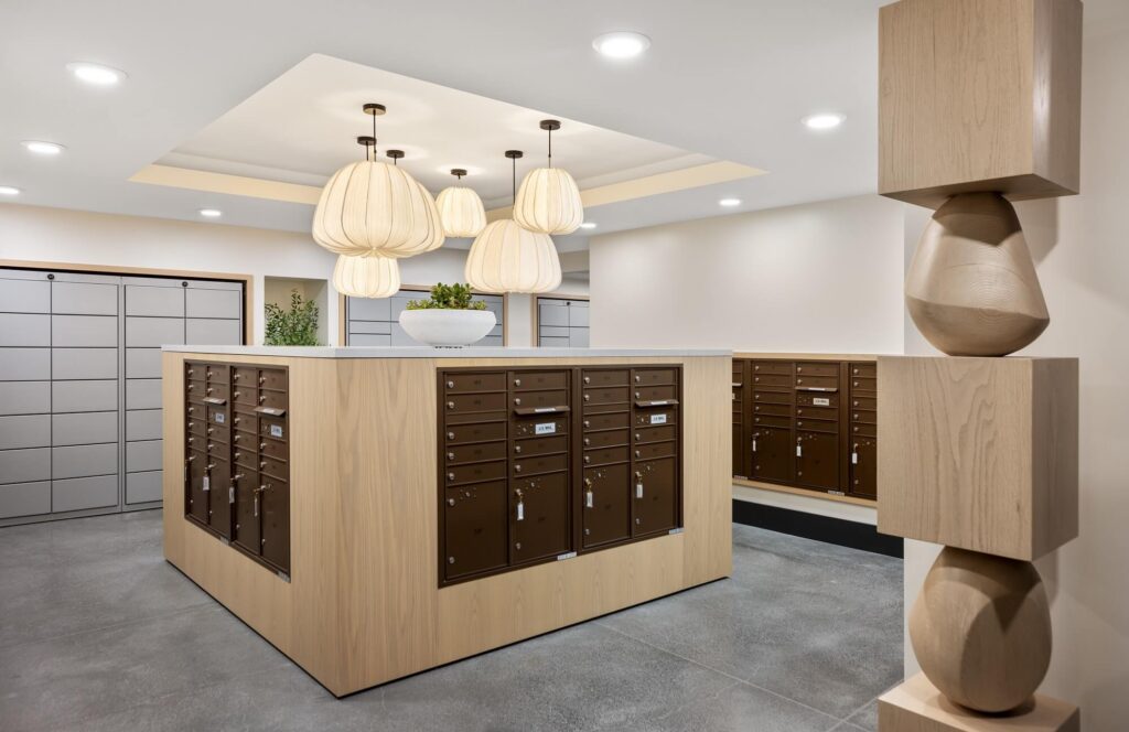 A modern mailroom with wood-accented mailboxes, decorative hanging lights, gray tile floors, and geometric wood sculptures on the right. A large white bowl with greenery sits on the mailbox counter.