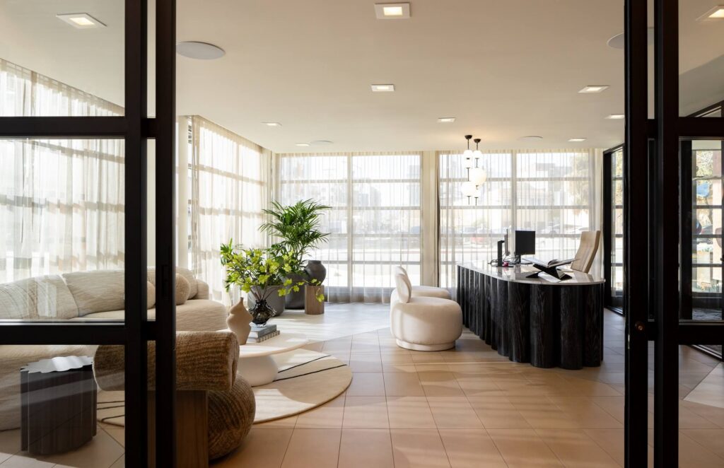 Modern office with large windows, black desk with a computer, white chairs, a beige sofa, several potted plants, round rugs, and soft natural light filling the airy, elegant space.