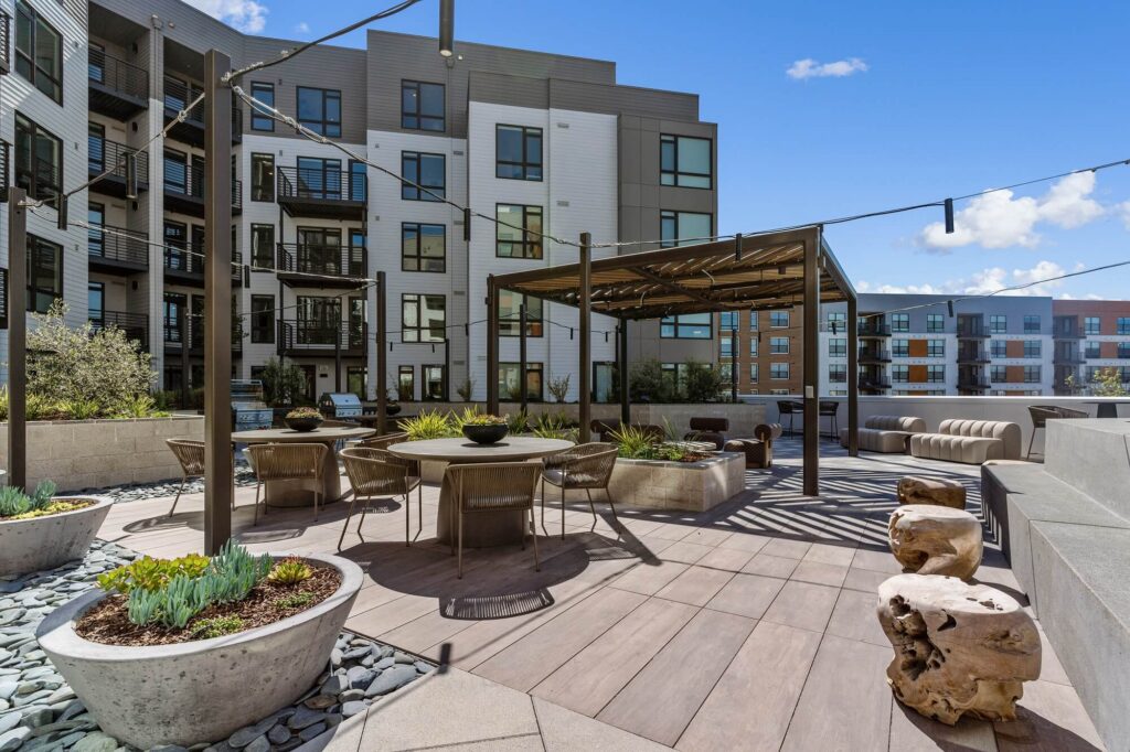 Modern apartment courtyard with string lights, outdoor seating, wooden tables, planters with succulents, and a covered pergola. Multi-story apartment buildings surround the patio under a sunny sky with scattered clouds.