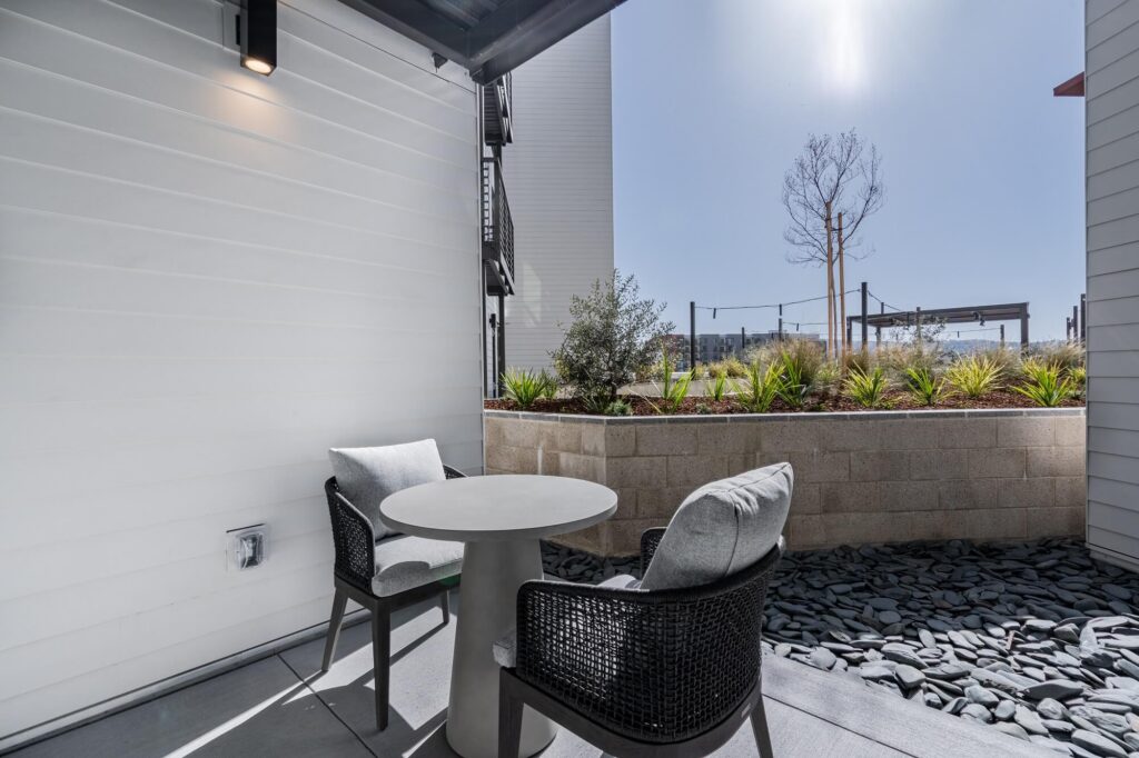 A small patio with a round table and two chairs on a stone floor, next to a raised garden bed with plants, under a clear blue sky.
