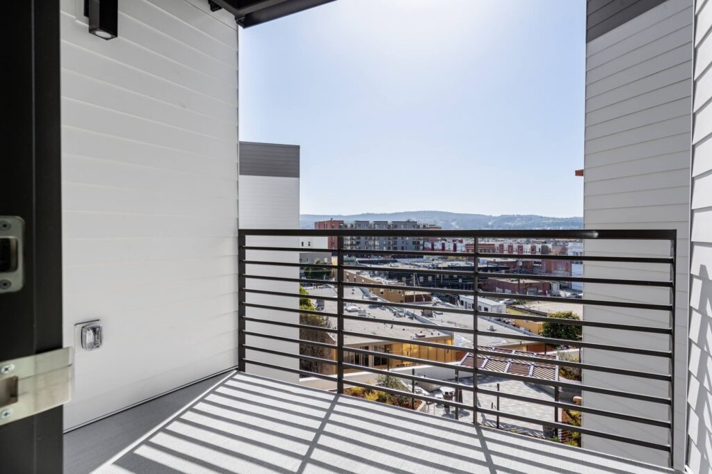 Small balcony with a black metal railing overlooks city buildings and distant hills under a clear sky. White walls and floor with sunlight casting shadows on the balcony surface.