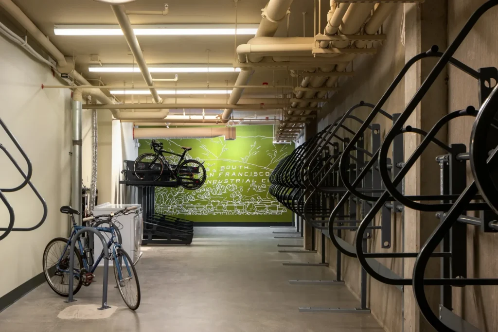 A modern indoor bike storage room with empty vertical racks on the right, two bikes parked, and a green mural on the back wall featuring a map and industrial design.