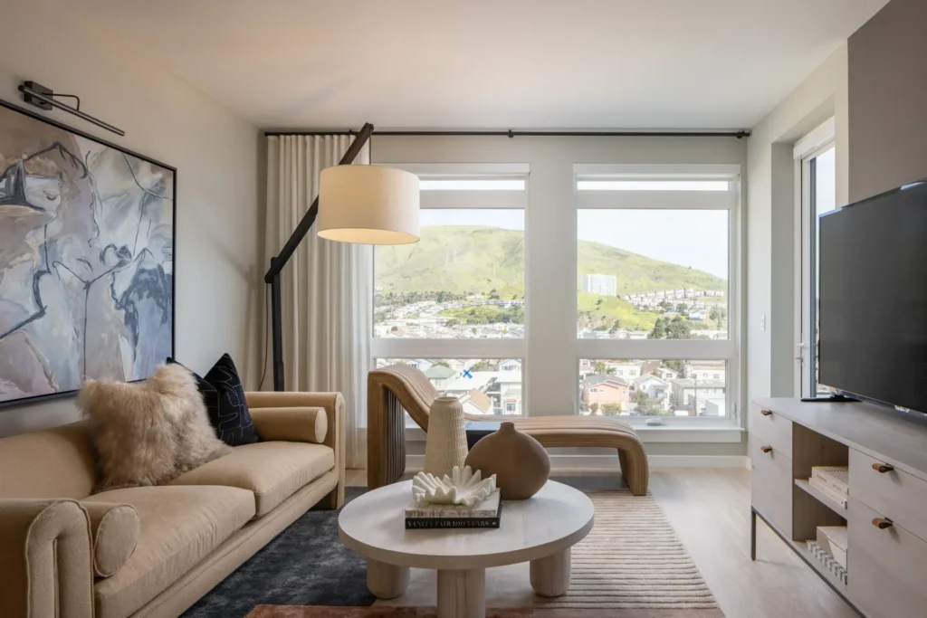 Modern living room with beige sofa, abstract art, round coffee table, sculptural decor, large window with hill view, and TV on a light wood stand. Neutral tones and natural light create a cozy, inviting atmosphere.