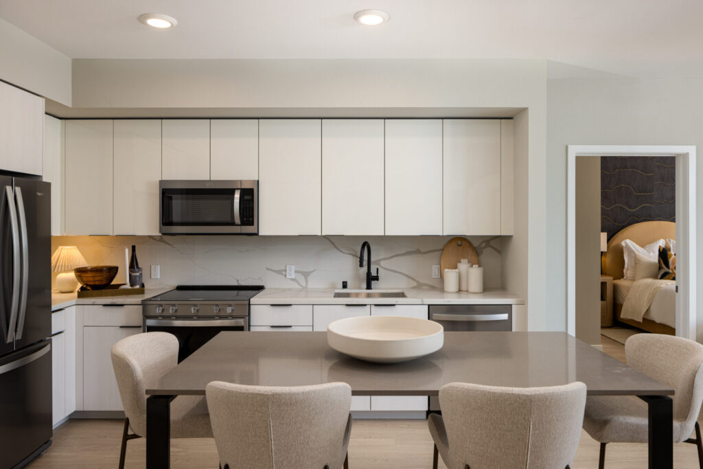 Modern kitchen with white cabinets, stainless steel appliances, gray countertop, and a dining table with four beige chairs. A door leads to a bedroom with a neatly made bed visible in the background.