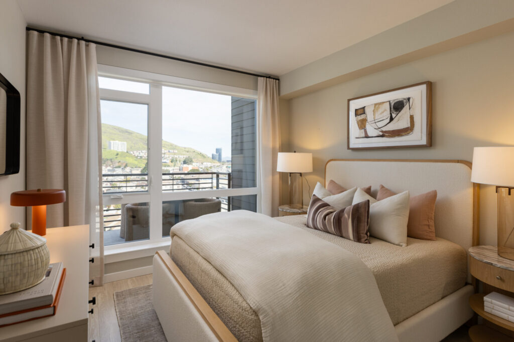 A cozy modern bedroom with a neatly made bed, abstract artwork above the headboard, two bedside tables with lamps, large window, curtains, and a city view with hills outside.