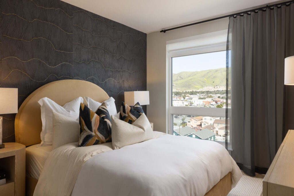 Modern bedroom with a large round headboard, white bedding, patterned pillows, two bedside lamps, dark accent wall, and a window showing a scenic hill and town view. Natural light fills the room.