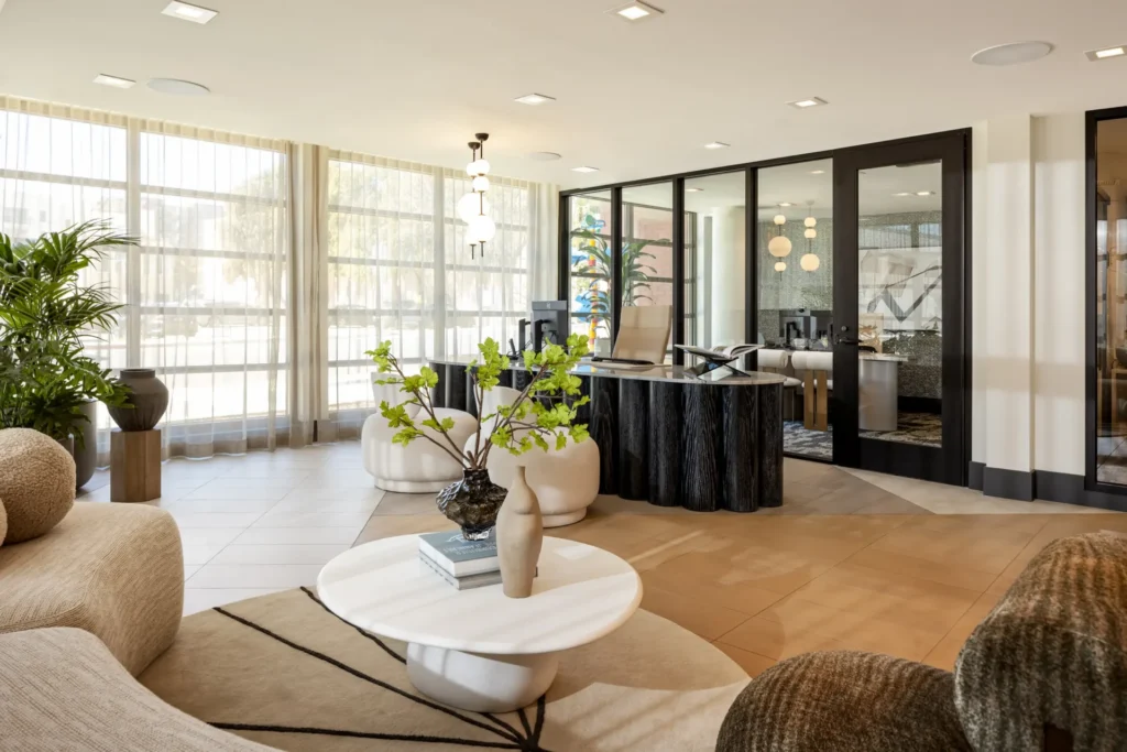 Modern lobby with large windows, light curtains, a round white coffee table with vases and books, beige seating, plants, and a reception desk with black accents in front of glass offices.