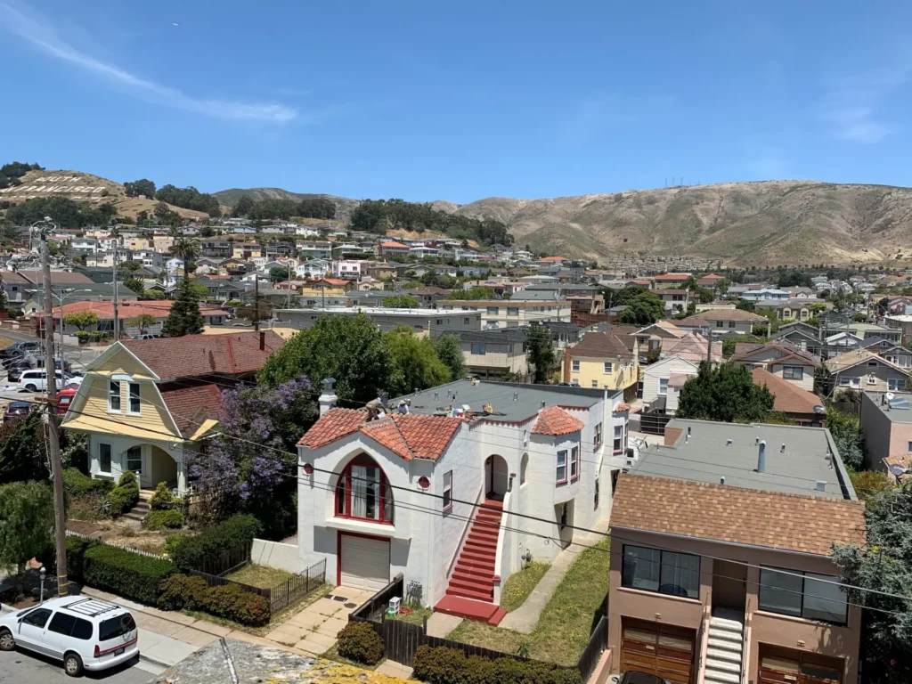 A residential neighborhood with houses, trees, and parked cars, set against rolling hills under a clear blue sky.