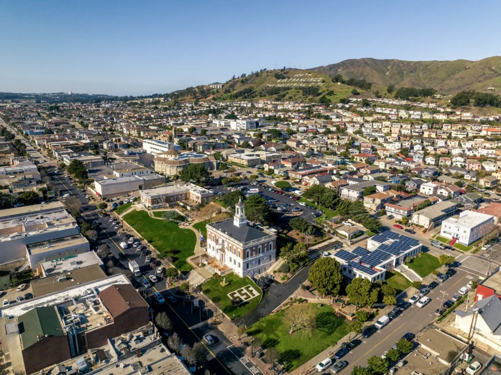 Aerial view of a suburban neighborhood with houses, a historic building with a clock tower, green lawns, and hills in the background under a clear blue sky.