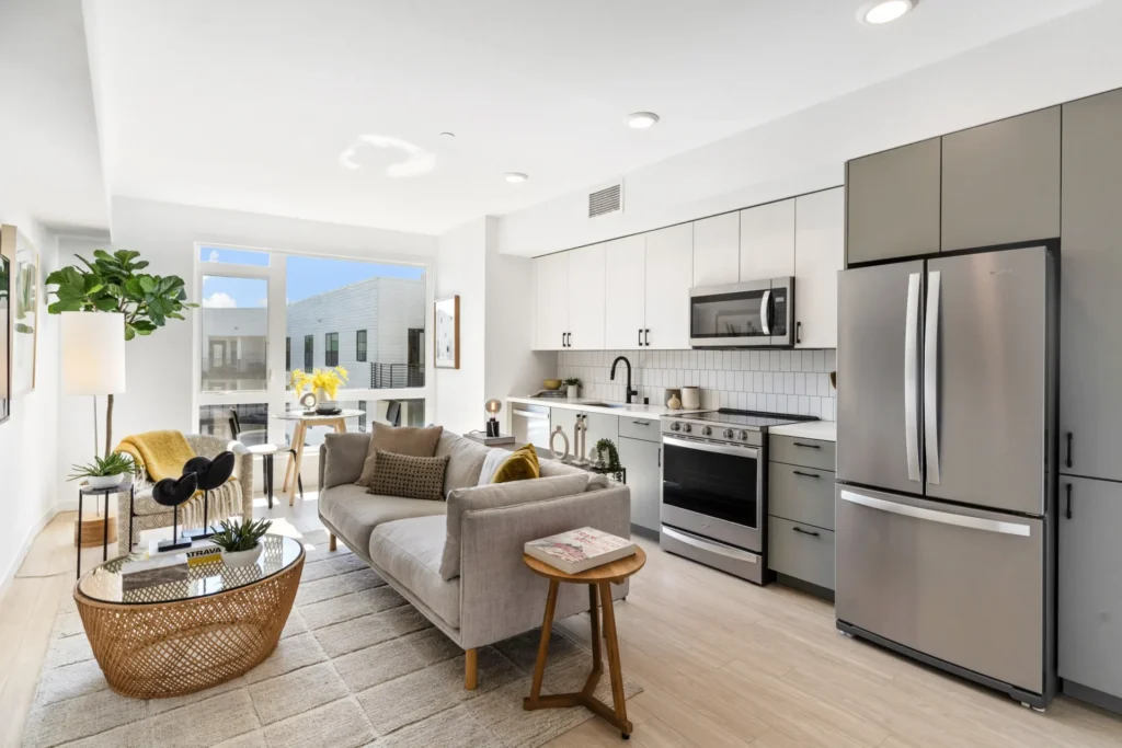 Modern apartment with open kitchen and living area, stainless steel appliances, light cabinets, a gray sofa, coffee table, and large window letting in natural light. Neutral decor and plants create a cozy, inviting atmosphere.