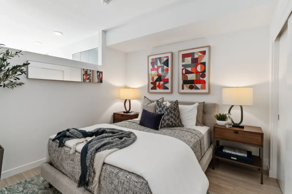 A modern bedroom with a gray upholstered bed, white bedding, throw blankets, and pillows. Two bedside tables with lamps, abstract art above the bed, a plant, and a wall mirror complete the decor.