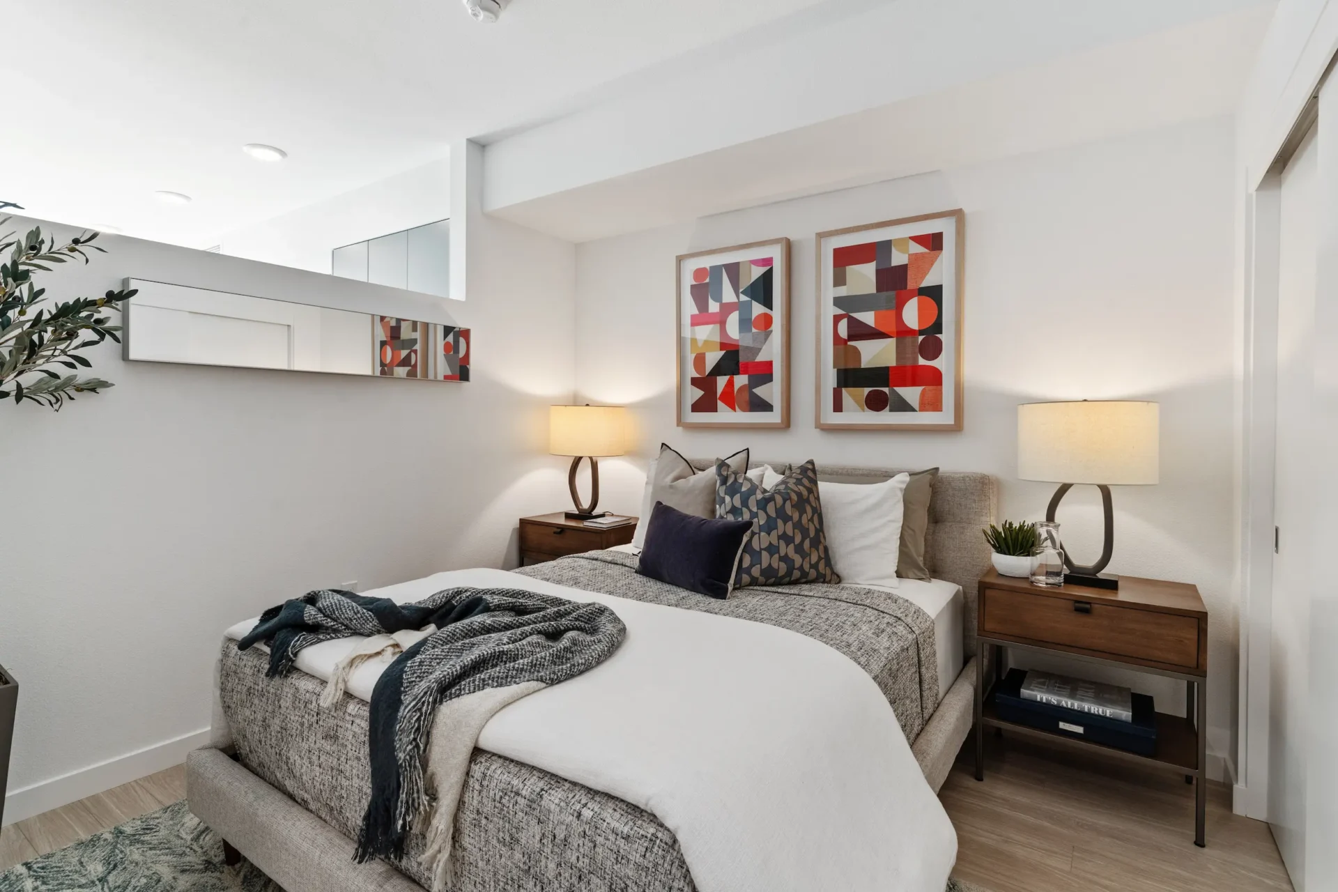 A modern bedroom with a gray upholstered bed, white bedding, throw blankets, and pillows. Two bedside tables with lamps, abstract art above the bed, a plant, and a wall mirror complete the decor.
