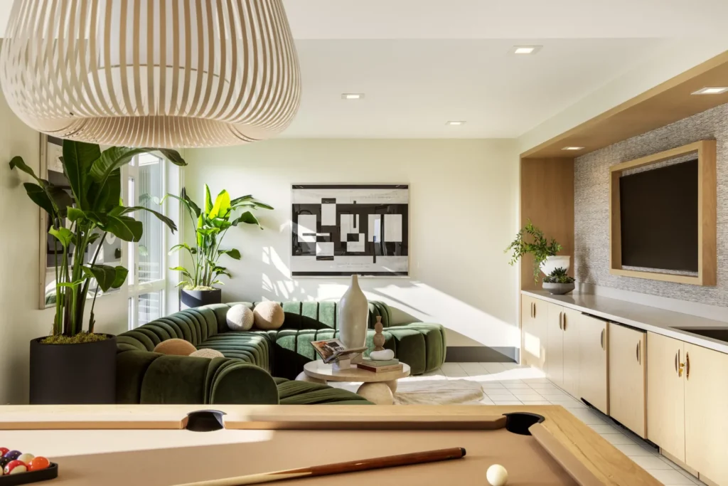 Modern living room with green sectional sofa, large plants, a wall-mounted TV, abstract artwork, and a pool table in the foreground under a white pendant light. Sunlight streams through windows, creating a bright, inviting space.