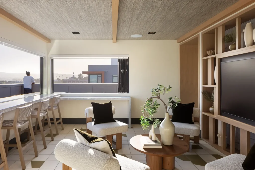Modern living room with beige chairs, a round wood coffee table, built-in shelving, and large windows; a person stands outside on a balcony overlooking city buildings in the distance.