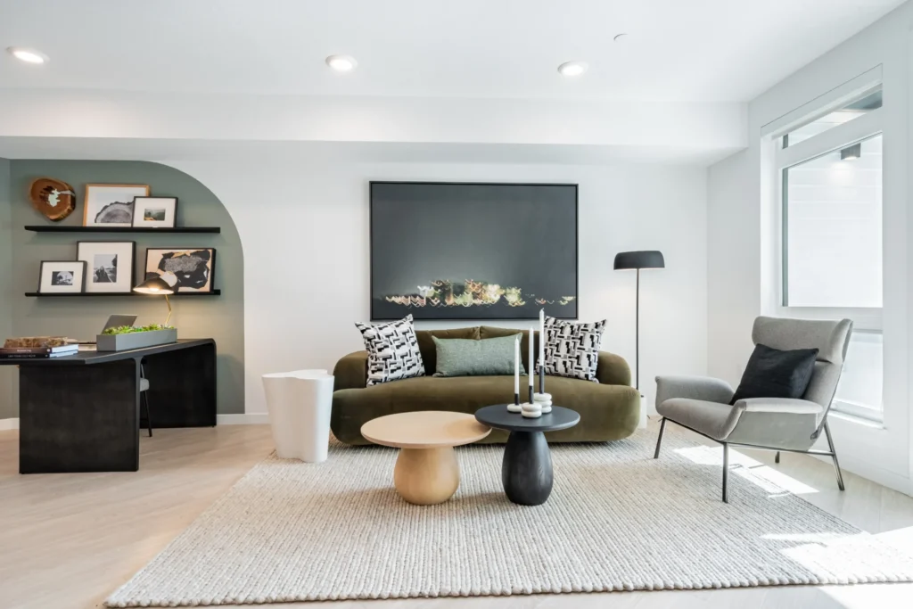 Modern living room with green sofa, gray armchair, two round coffee tables, large dark artwork, floor lamp, textured rug, and a workspace with shelves and decor in the background. Bright natural light from large windows.